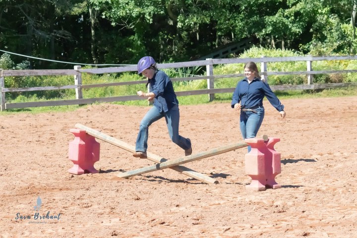 Two people in casual attire balancing on crossed beams in a sandy arena.