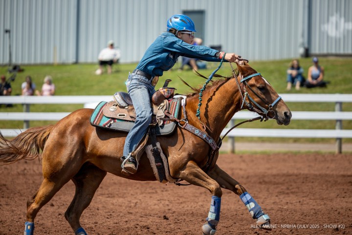 Person in blue helmet and denim riding a galloping horse in a dirt arena.