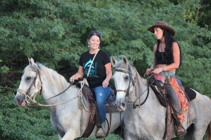 Two women riding horses in a forest, one smiling, both wearing casual outfits.