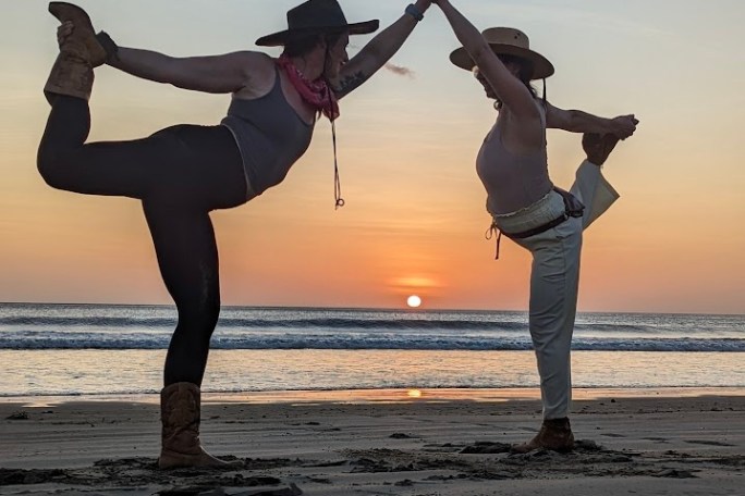 Two people in yoga poses on a beach at sunset, wearing hats and facing each other.