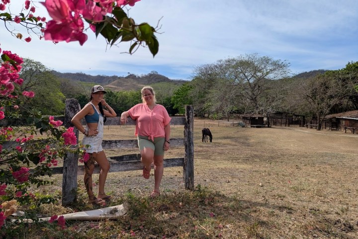 Two people by a rustic fence with pink flowers; a horse grazes in the background under a partly cloudy sky.