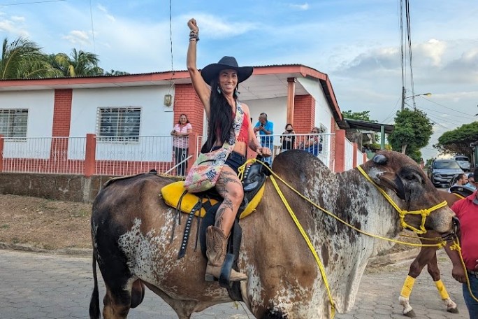 Person in a cowboy hat riding a decorated bull on a street, raising an arm in a festive gesture.