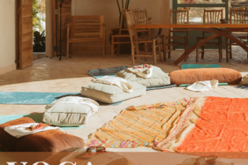 Yoga mats and cushions in a serene, sunlit room with wooden chairs and plants.